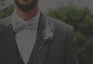 A man in a dark suit and vest with a white dress shirt and striped bow tie stands as if captured in an oil painting. He has a floral boutonniere on his lapel, featuring leaves and white feathers, set against the blurred greenery backdrop.
