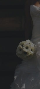 The bride, reminiscent of an exquisite oil painting, holds a bouquet of white flowers with dark centers against a shadowy background. The focus captures the flowers and the intricate Meissner-like fabric detail of the dress.