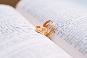 Two gold wedding rings are nestled between the pages of an open book, casting a heart-shaped shadow on the text—a scene reminiscent of a meissner painting. The focus is on the rings, with the surrounding text gently blurred like soft strokes in a piece of art.