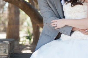 Close-up of a couple on their wedding day, as if captured in a delicate oil painting. The bride in her white dress and the groom in his gray suit sit intimately, with her hand resting on his arm. In this Meissner-like scene, blurred trees softly frame their love-filled moment.