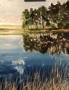 A tranquil lakeside scene with calm water reflecting a line of trees on the opposite shore. Tall grass in the foreground adds depth to the landscape, while the sky above is clear and bright.