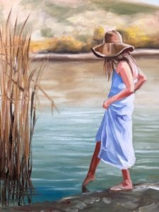 A painting of a girl in a light blue dress and wide-brimmed hat stands at the edge of a serene body of water. Testing the waters with her barefoot, she is surrounded by reeds. The blurred landscape in warm tones adds depth to her quiet contemplation.