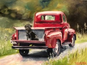 A red vintage truck is parked on a grassy path. An old black dog sits in the trucks open bed, looking towards the viewer. The background is a blurred mix of green and brown, suggesting a rural setting.