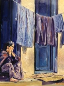 A woman sits on a step outside a doorway, resting her head in her hands. Next to her, colorful clothes hang on a line, drying in the sunlight. The scene conveys a sense of contemplation and everyday life.