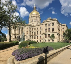 A neoclassical building with a golden dome under a blue sky is surrounded by trees. A statue stands in front, while a garden with purple flowers lines the path, often bustling during events. The shadow of a person taking a photo is visible on the path.