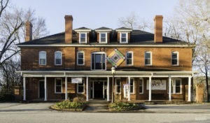 A large brick building with a covered porch, multiple chimneys, and dormer windows hosts vibrant events year-round. A colorful quilt pattern adorns the entrance, beneath signs that read Open and Jack. Bare trees and an overcast sky create a serene backdrop for the lively shows inside.