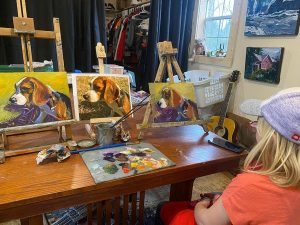 A person with long blonde hair and a purple beanie sits at a wooden table with art supplies and a palette. Three dog paintings inspired by Reas Aquaduct Waterfall are on easels. A guitar and clothes are in the background.