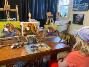 A person wearing a cap and glasses sits at a table with three paintings of a beagle on easels. Art supplies and paint tubes are spread across the table. Paintings by Michael Meissner and a guitar adorn the background, creating an inspiring creative space.