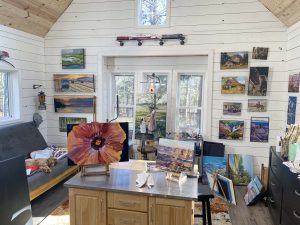 A cozy art studio resembling Double Gap Farm, adorned with various paintings on white walls featuring landscapes and florals. A central table displays a large flower painting. To the left, a dog sleeps on a gray couch as natural light streams through the large windows at the back.