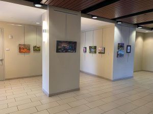 The art gallery hallway, often hosting captivating shows, showcases various colorful paintings on light beige walls with overhead lighting. Wooden ceiling panels and tiled floors enhance the open, well-lit space, creating an inviting atmosphere for viewing artwork.