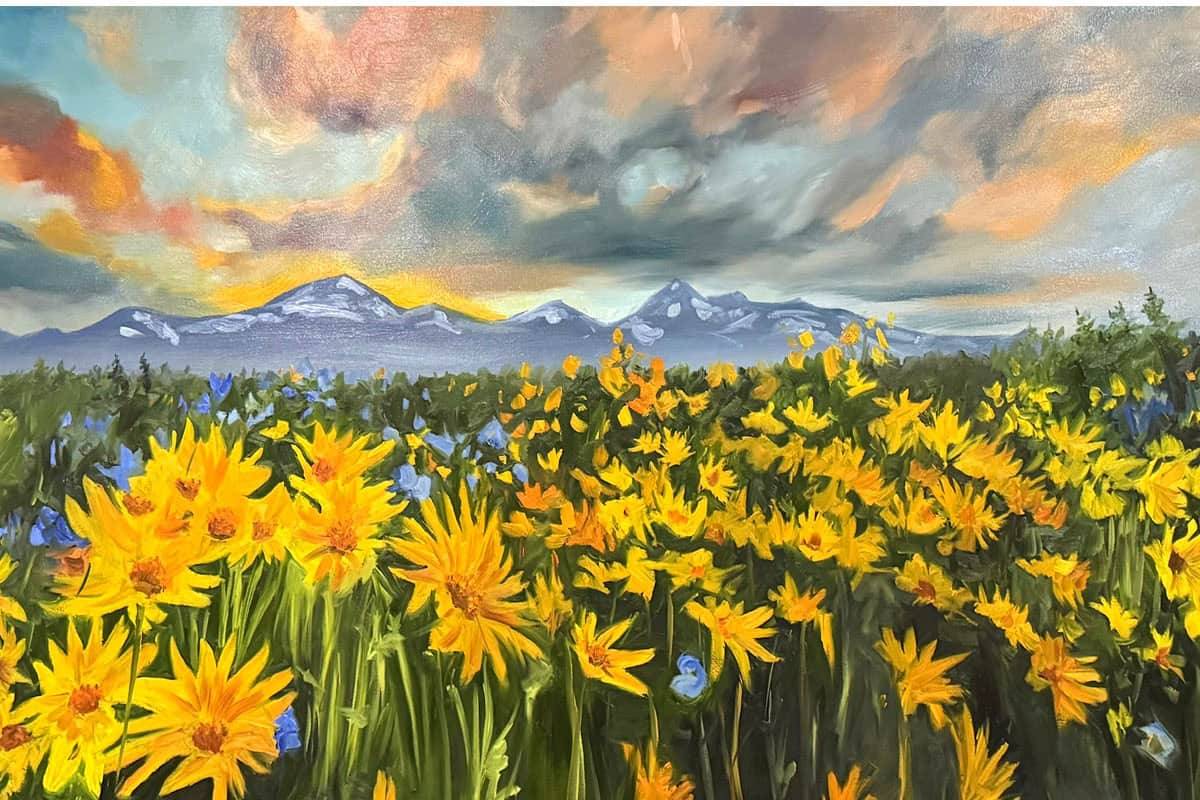 A vibrant field of yellow wildflowers with scattered blue flowers stretches toward distant snow-capped mountains under a dramatic, colorful sky at sunset.